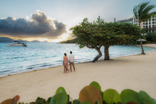 Couple standing on a sunset beach with blue ocean watching a catamaran in the distance