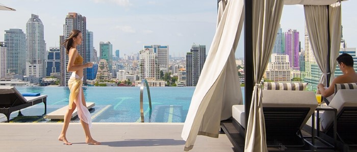 Woman walking past a rooftop pool with a view of the city in the background and a man in a curtained cabana