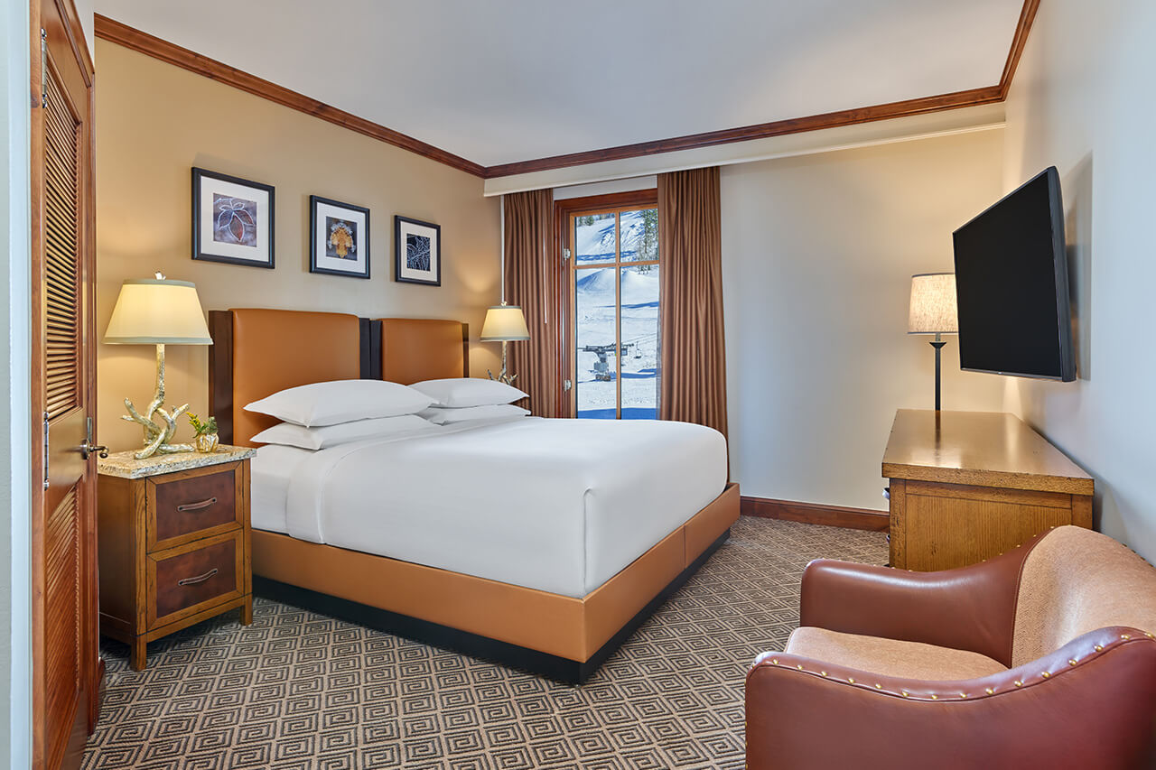 King bed in resort guestroom with a warm leather frame, white linens, and a snowy landscape outside the window