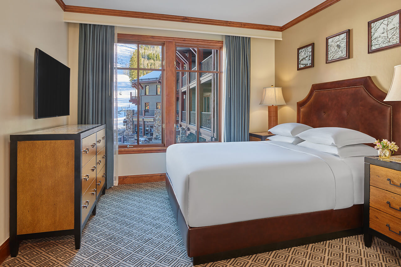 Resort guest bedroom with a king bed with leather frame, white linens, and gray plaid carpet.