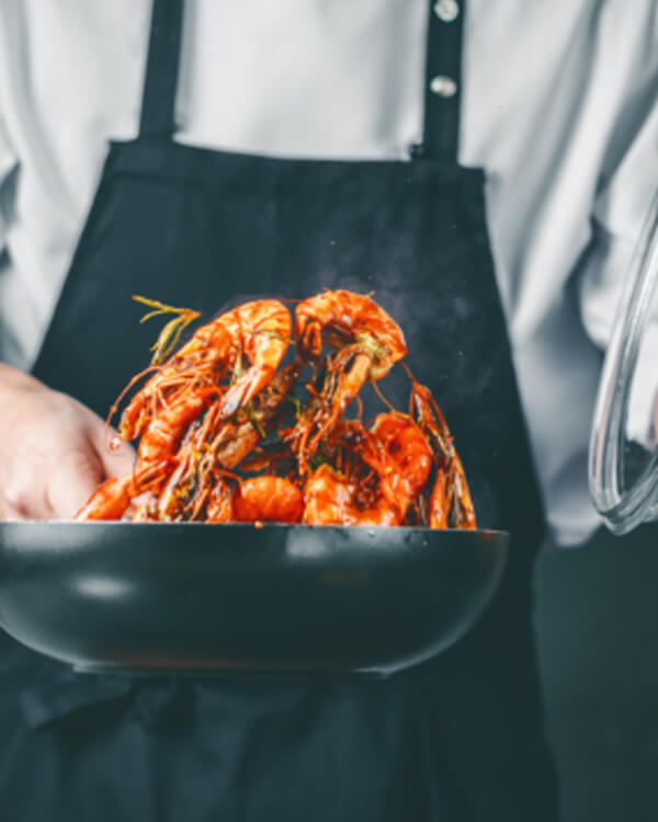 Chef in a white coat and black apron tossing bright red shrimp in a black pan.