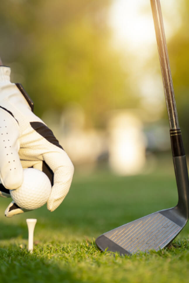 Hand in golf glove placing a ball on the tee with a golf club ready.