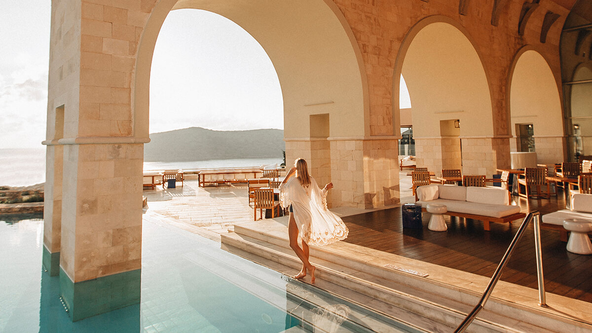 Woman in a white swim cover-up looking at the water from the walk-out pool under the arched columns.