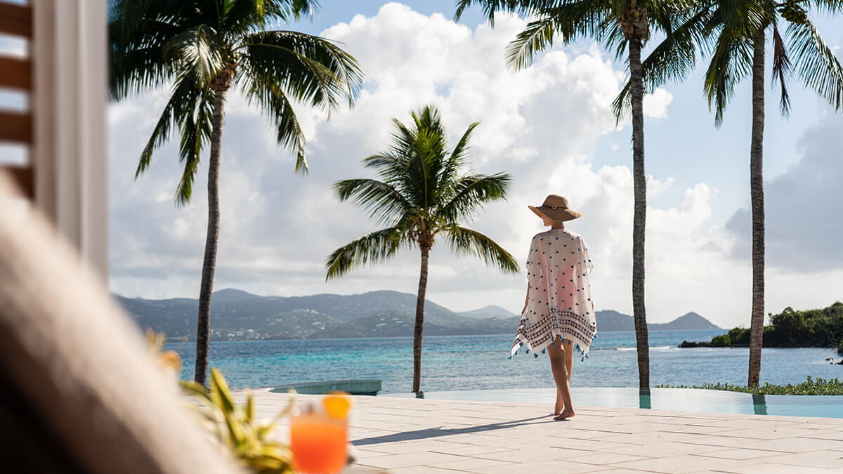 Woman in a white with black polka dot cover up looking at the water from the palm tree lined deck