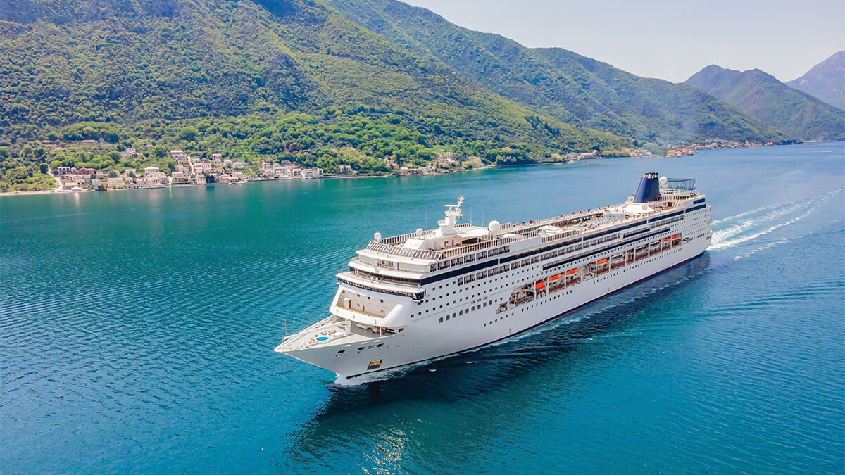 Cruise ship at sea cruising past a resort nestled in tree covered mountains. 