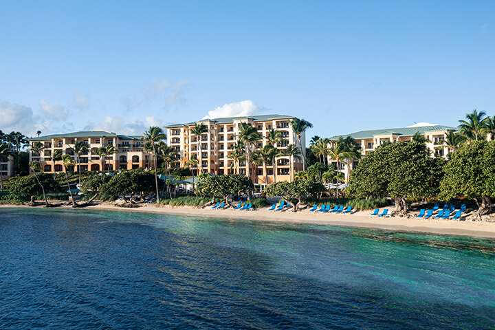 Ritz Carlton Club, St. Thomas exterior aerial image from the ocean looking at the resort and beach lined with blue lounge chairs and palm trees