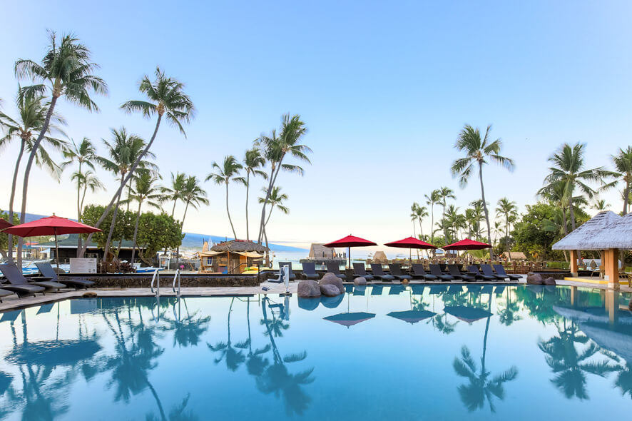 Tropical resort pool overlooking the ocean with palm trees reflecting on the pool and red umbrellas with lounge chairs.