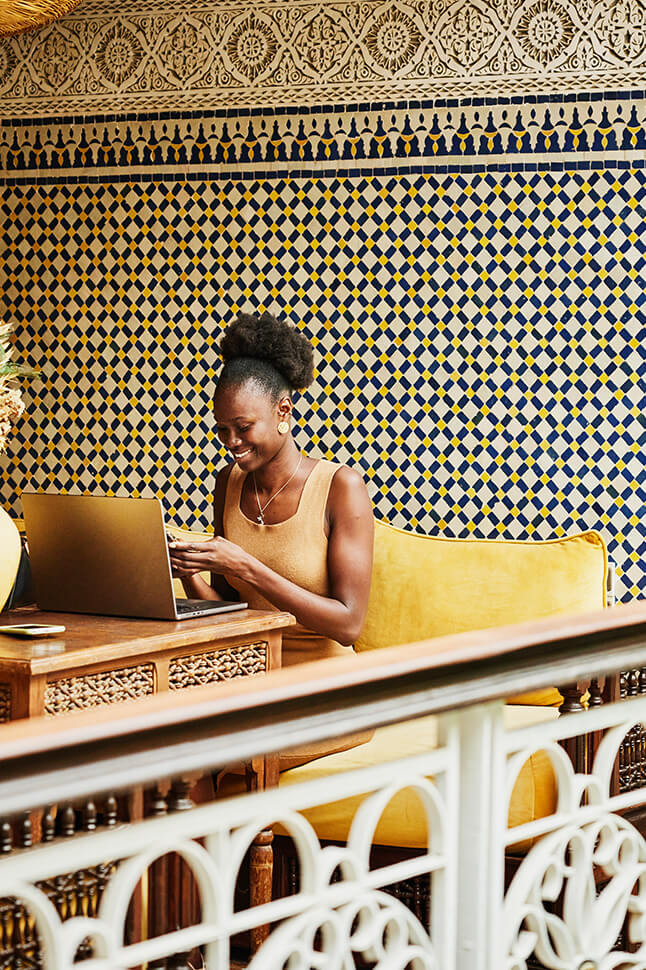 Woman wearing a yellow dress sitting on a yellow couch working on her computer and looking at a mobile phone.