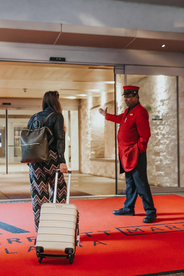 Door staff at the front entry of a Marriott location welcoming a traveler with a roller-bag.