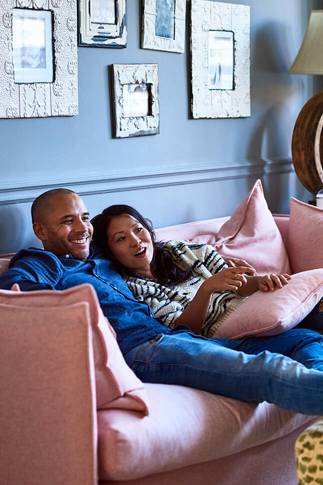 Couple sitting on a light pink couch with fluffy pink throw pillow smiling and laughing.