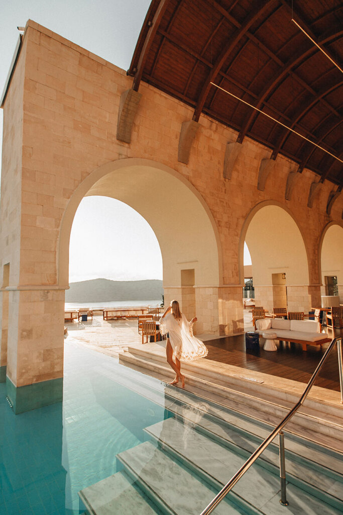 Woman standing under arches in a walk in pool looking at the ocean and mountain view