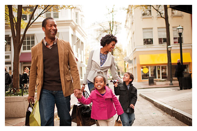 Family of four carrying shopping bags walking on a cobblestone street in a quaint setting.