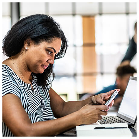 Woman looking at her mobile device with her laptop computer open and paper and pen on the table. 