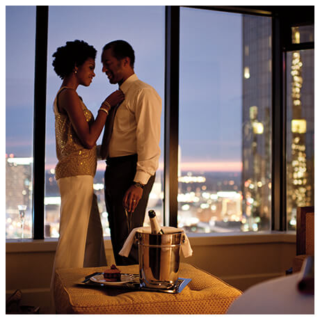 Couple standing together in front of a window with city lights in the background and a champagne set up on the table.