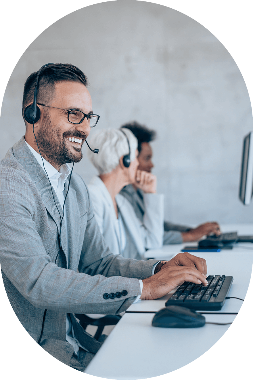 Man in an office setting with headphones and glasses on while typing on a keyboard with co-workers in the background